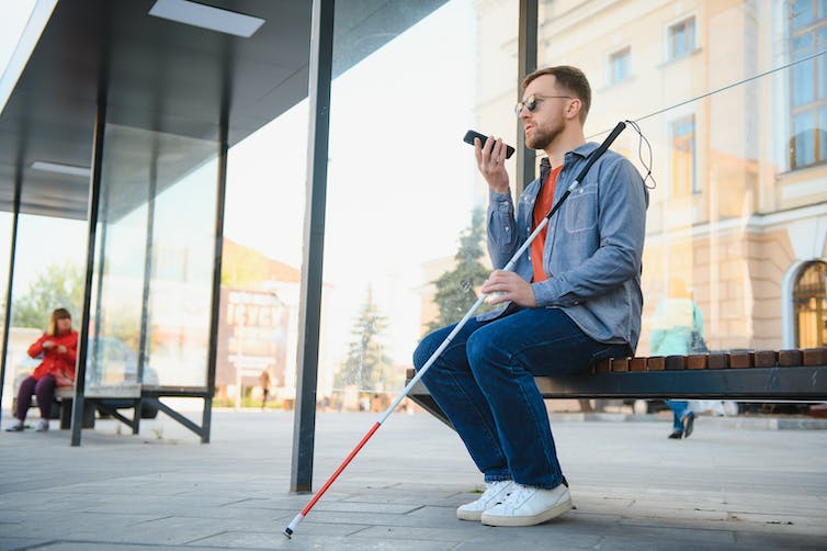 a man sitting at a bus stop holding a cane and talking on his cell phone